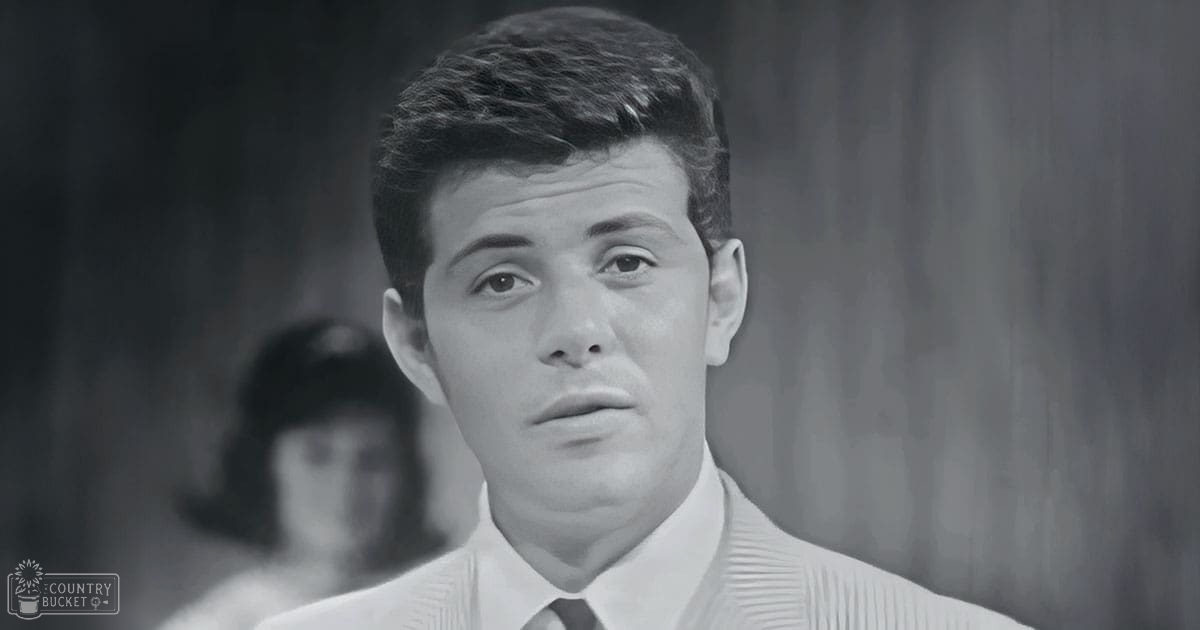 Young adult male in a formal 1950s-60s attire stands indoors with a child or young woman, possibly female, in the background.