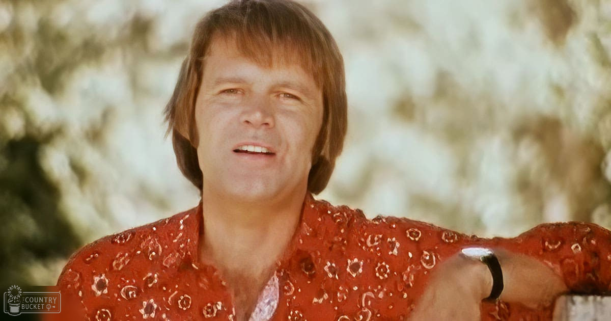 A middle-aged adult male in a relaxed, long-sleeved collared shirt with floral or paisley patterns stands outdoors under bright daylight.