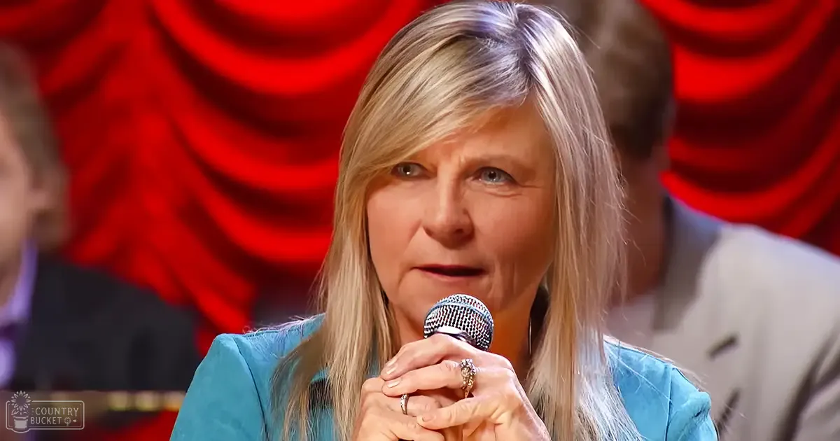 A middle-aged woman wearing a teal blazer speaks into a microphone in what appears to be a professional studio setting, with bright lighting and red curtains visible.