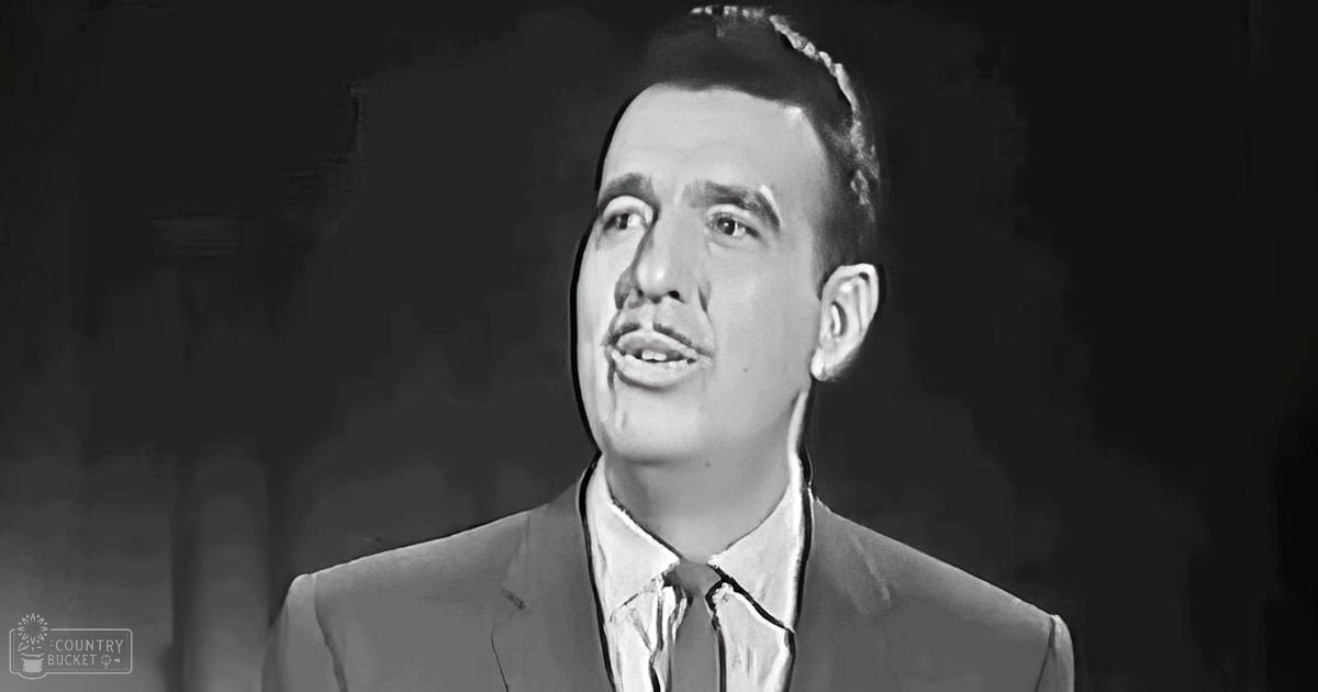 A man in an indoor setting wearing a suit jacket with a notched lapel and a dress shirt with the top button undone, possibly from earlier decades.