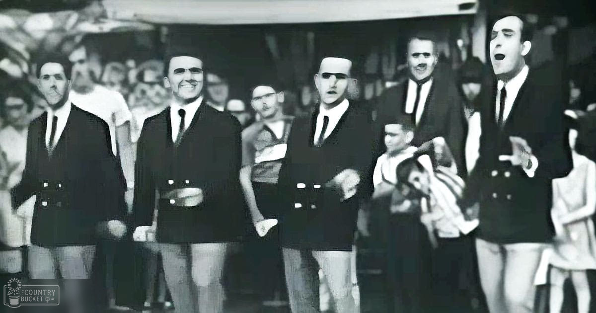 A group of adult male singers in dark double-breasted suits performing indoors, surrounded by an audience dressed casually.