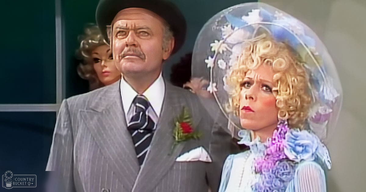 An adult male in a gray pinstripe suit, white shirt, striped tie, black hat with boutonniere, and an adult female wearing a white blouse or dress with blue/purple floral patterns, large hoop earrings, and clear plastic dome-like headpiece decorated with flowers, standing indoors near a window suggesting daytime lighting.