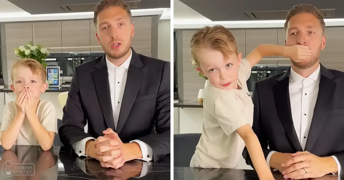 A middle-aged adult male in a dark suit jacket sits indoors at a modern kitchen counter with a child wearing a light-colored t-shirt and bracelet standing nearby.