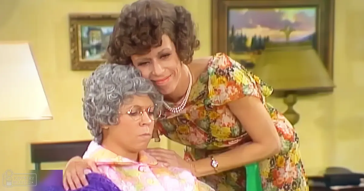 Indoor scene with older adult female and younger adult female wearing floral dresses indoors; furniture includes a lamp and framed paintings depicting an outdoor landscape and room interior.