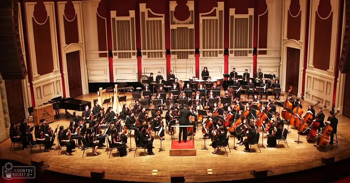 An adult male conductor leading an orchestra on a grand stage with ornate architectural details and red curtains in an indoor evening setting.