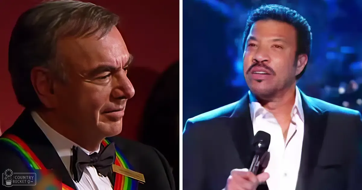Middle-aged male in a formal tuxedo with a colorful sash, holding a microphone beside another adult male in a black suit jacket over a white shirt at a dimly lit stage setting with blue and red lighting.