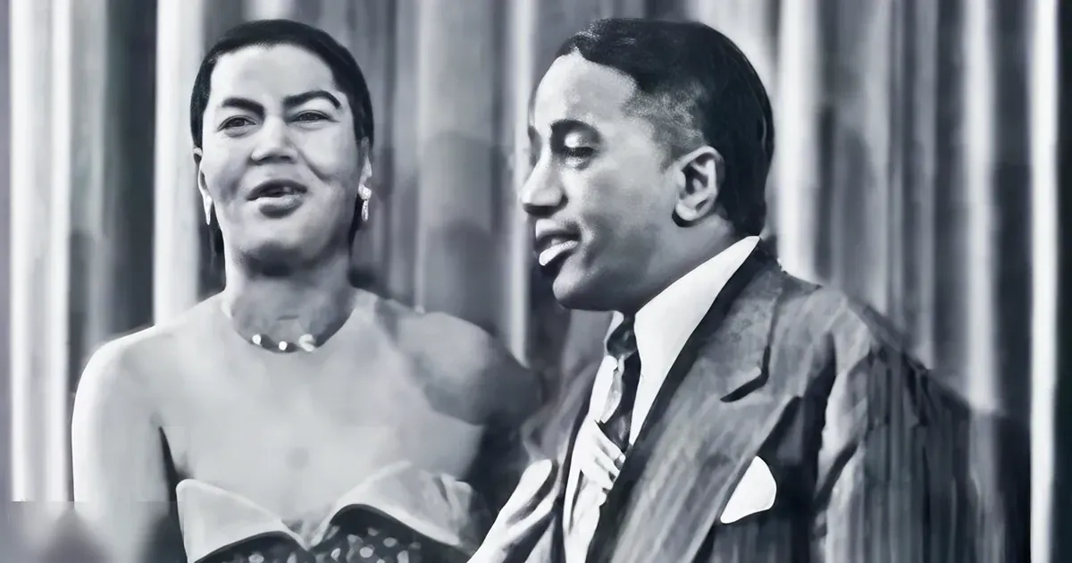 Adult female in a formal dress with patterned skirt and dangling earrings, alongside an adult male in vintage formal attire with lapel and pocket square, set against vertical blinds indoors.