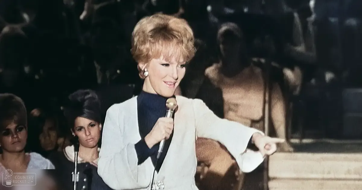 An adult female performer in a white blazer with bell sleeves and black turtleneck, holding a microphone on stage at an indoor event.