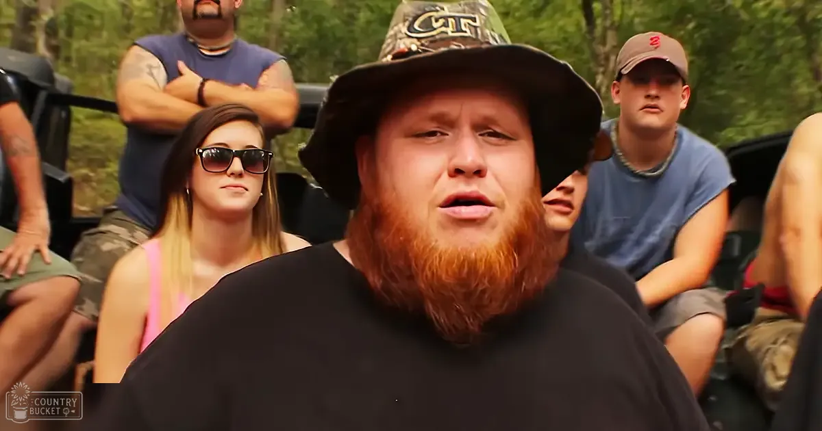 Young adult male in a bucket hat and black shirt, surrounded by other individuals outdoors in a wooded area with greenery and natural lighting.