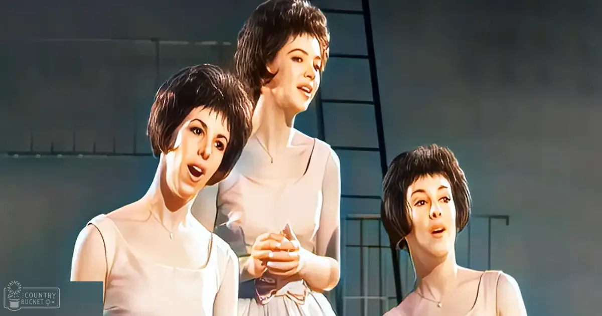 A theatrical performance scene indoors, possibly during a musical or play, with three young adult female performers in sleeveless white dresses with high necklines and fitted bodices against a simple dark backdrop.