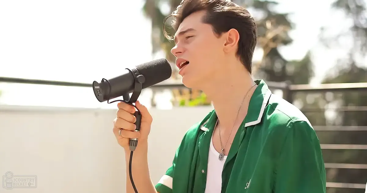 A young adult male wearing a green jacket with white trim, holding a microphone while singing outdoors in an urban setting.