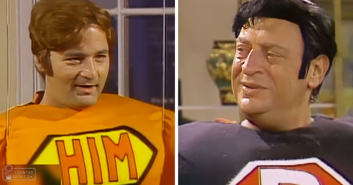 Two adult males dressed in 1970s-style T-shirts with shield logos, standing indoors near a decorative vase.
