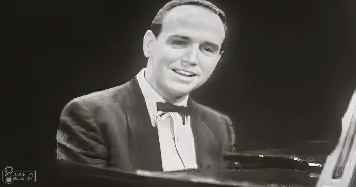 A man in a dark suit jacket with a light-colored shirt and bow tie sits at a piano, likely in a studio setting.
