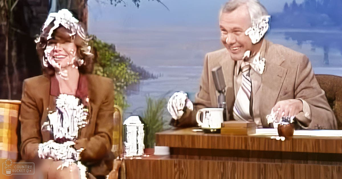 Two adults in a talk show setting, one wearing a brown blazer and patterned dress, the other in a light brown suit with decorative accessories; scenic background of trees and water viewed from an indoor studio.