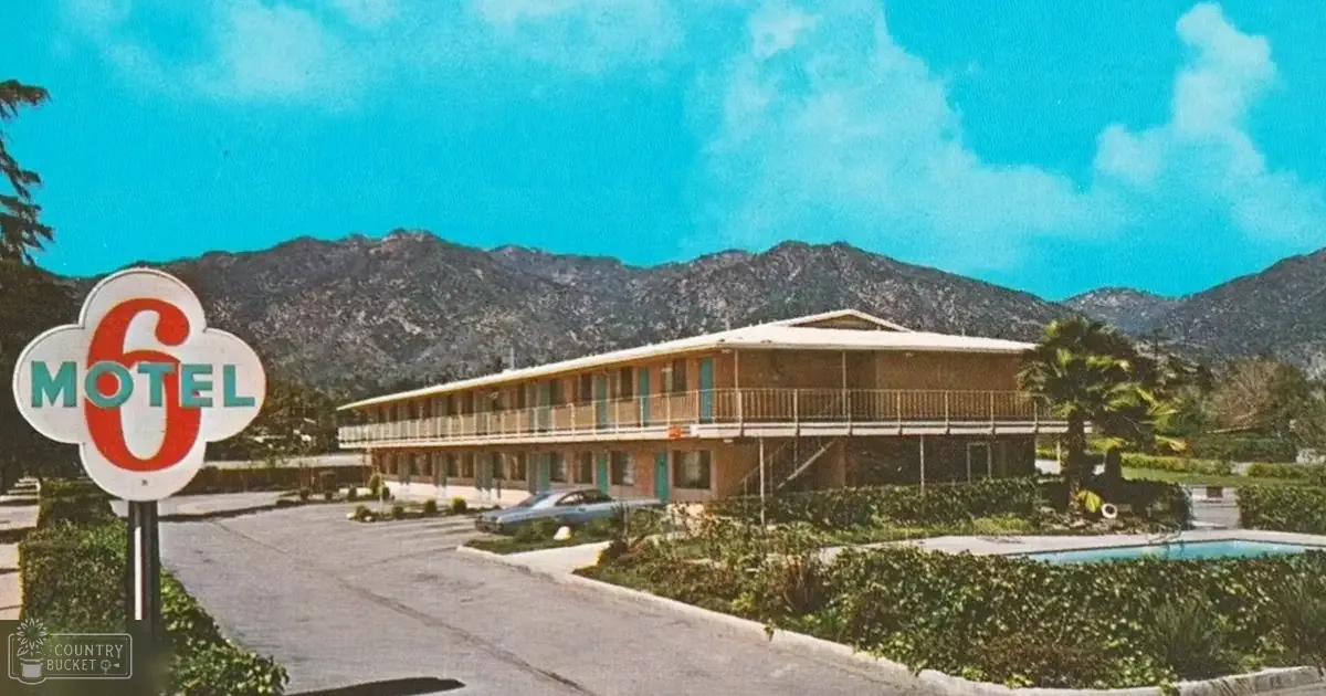 Motel 6 building with balconies, palm tree, shrubs, car parked near the entrance, clear blue skies and green vegetation surrounding it.
