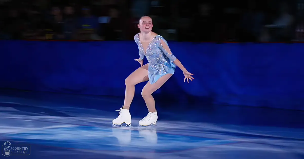 A young adult female figure skater in a blue sequined dress performs at an indoor ice rink with spectators seated behind barriers.