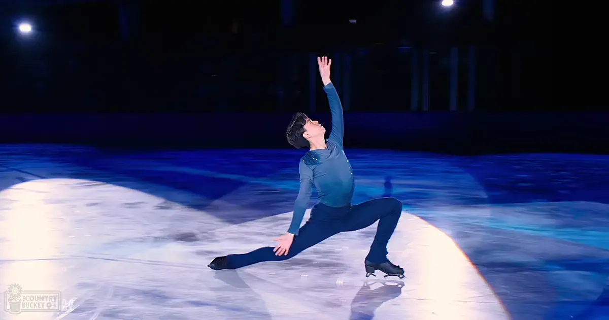 A young adult male figure skater in a long-sleeved dark teal figure skating costume on an indoor ice rink with spotlights illuminating the scene.