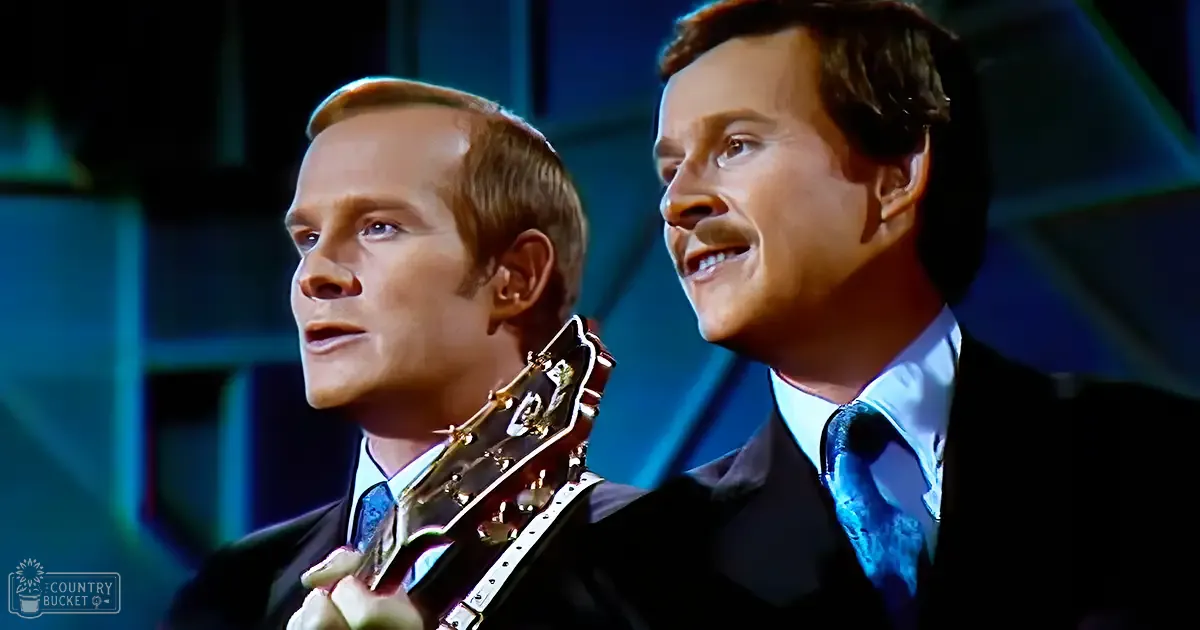 Two young adult males dressed in dark suits and light blue patterned ties perform on an indoor stage or studio with bright lighting, emphasizing their formal attire.