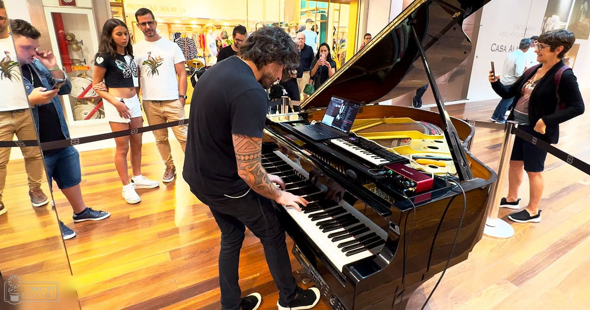 Adult male playing piano in an indoor shopping mall, wearing a patterned black t-shirt with dark pants and black sneakers.