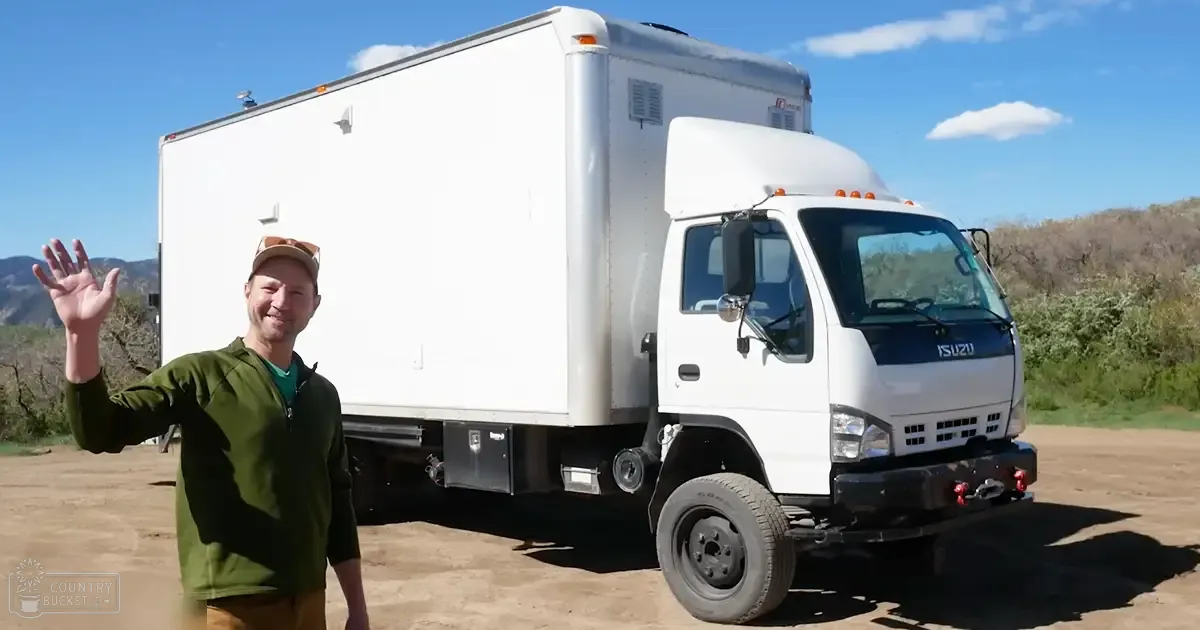 A middle-aged to older adult male wearing a green zip-up jacket and light-colored backward cap stands in front of a white Isuzu truck on dry grassy ground under clear blue sky with scattered clouds, suggesting late spring or summer.
