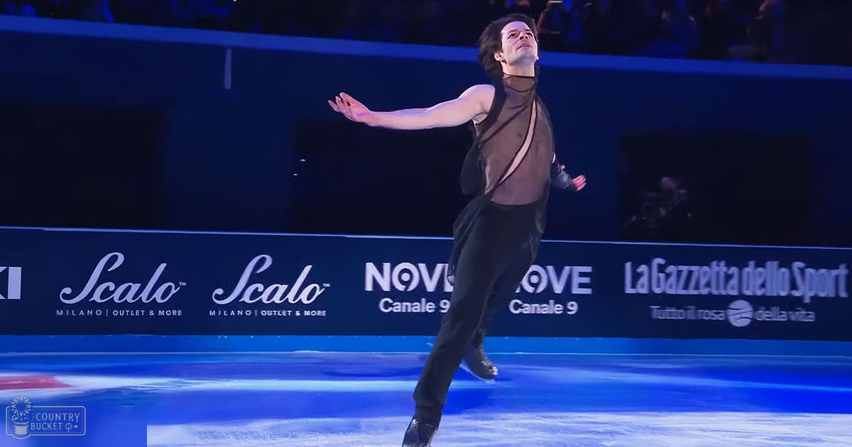 A young adult male figure skater performs at an indoor ice rink under artificial blue lighting. The skater wears a black outfit with sheer, flowing fabric draped over the shoulders and chest area.