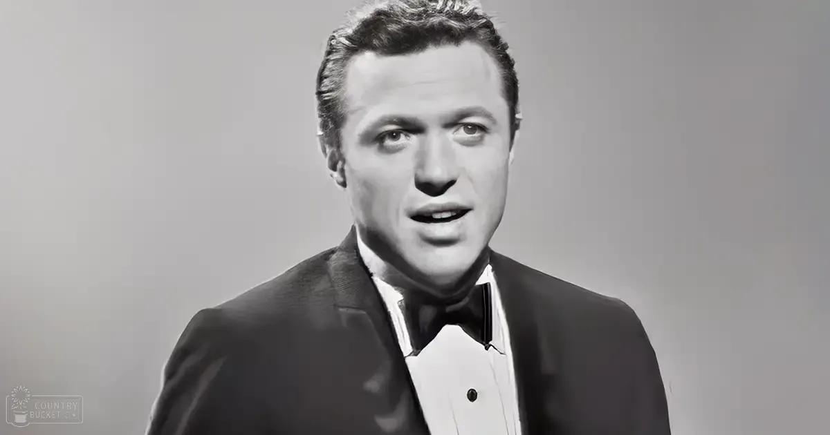 A middle-aged man in a formal suit with a bow tie, likely from the mid-20th century, stands against an indoor plain background.