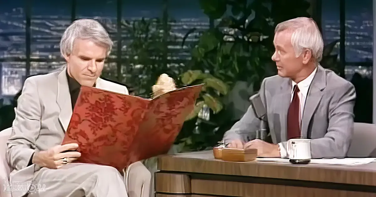 A man and an older adult male seated at a desk, discussing with open books on a talk show set. The man in the suit jacket holds the book while wearing a ring; the older man in gray attire sits beside him.