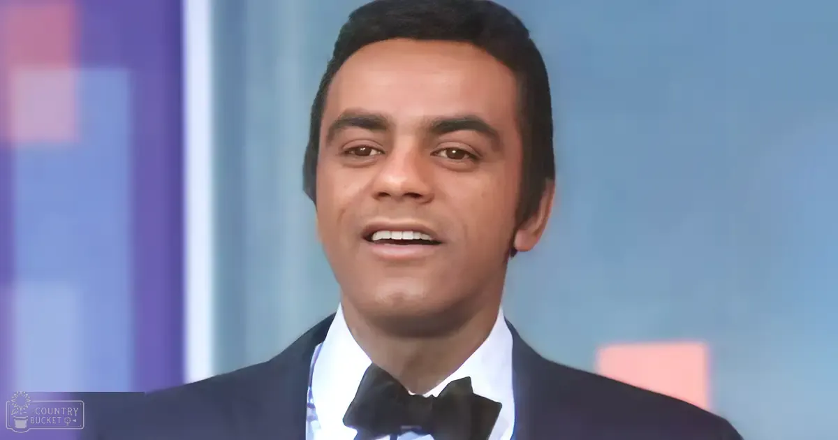A young adult male wearing a dark blue formal suit with a white shirt and black bow tie stands in an indoor studio setting.