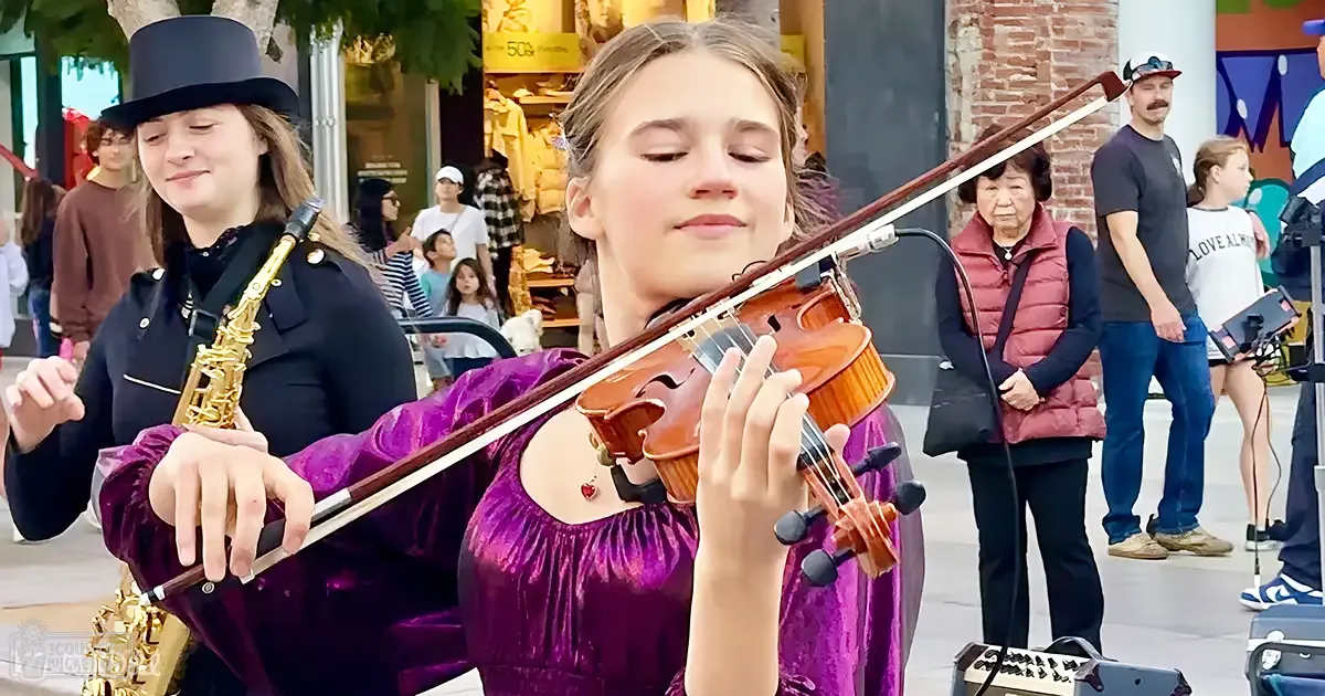 A young adult female plays the violin in a public urban space with various passersby and musical equipment around her.