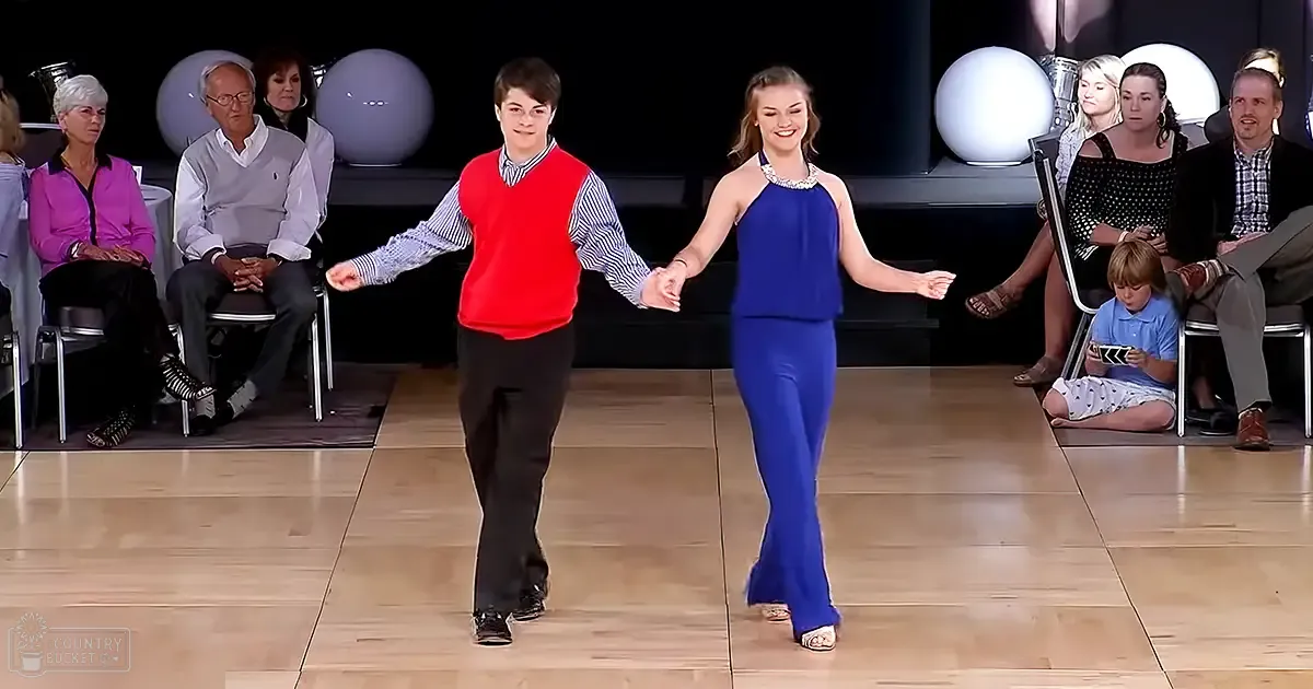 A dance duo performs in a studio setting with an audience seated around the perimeter. The performers are dressed in casual dance attire, while the audience includes adults and children in various outfits.