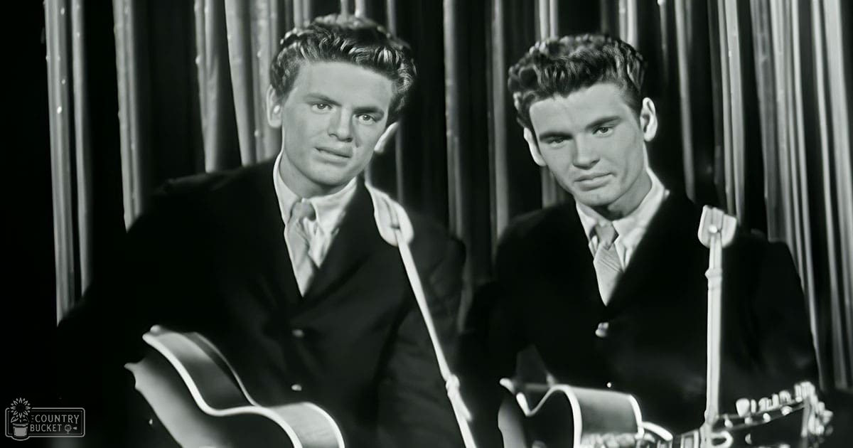 Two young adult males in a studio setting wearing formal mid-20th century suits with acoustic guitars and microphones, suggesting a musical performance or recording session.