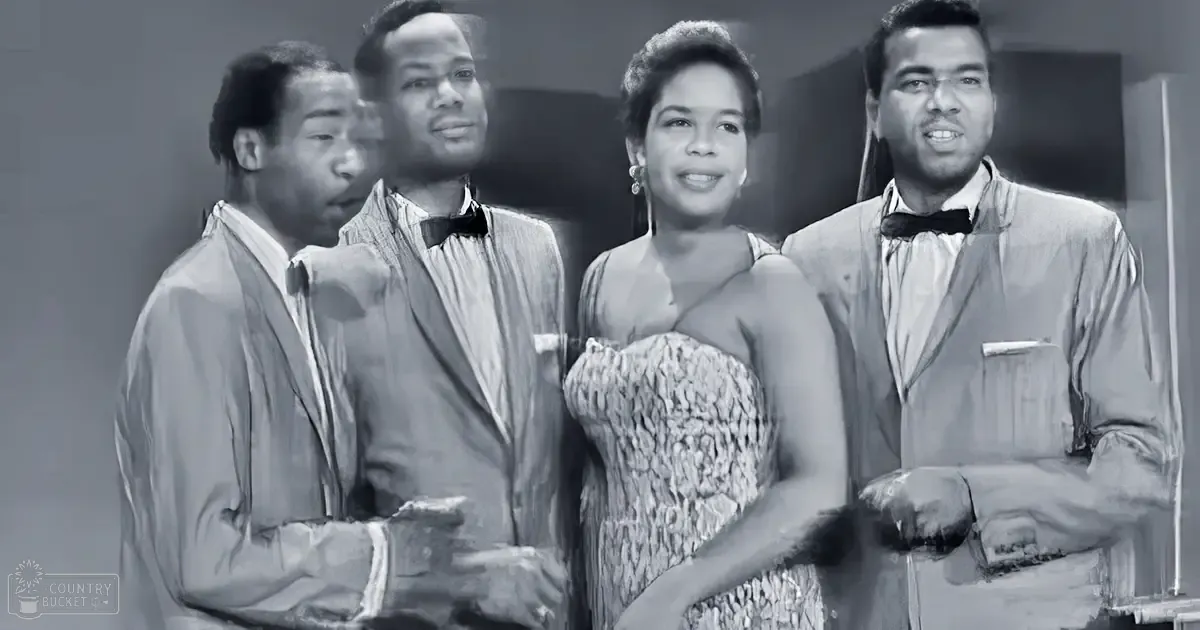 A group of performers in a studio setting; adult male in light-colored shirt and tie, middle-aged man in suit jacket over collared shirt with bow tie, adult female in sleeveless dress with textured pattern, young to middle-aged adult male in similar suit. Earrings partially visible on the female performer.