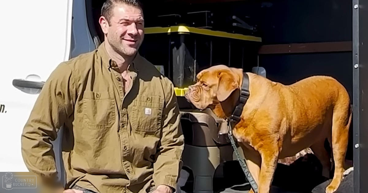 An adult male wearing an olive green button-up shirt with two chest pockets, standing outdoors with a light brown mastiff dog. The setting suggests a campsite or RV park with clear daylight and outdoor furniture.