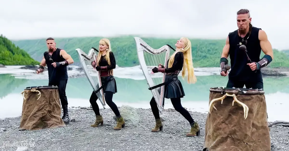 A group of young adults performing traditional music outdoors near water under an overcast sky.