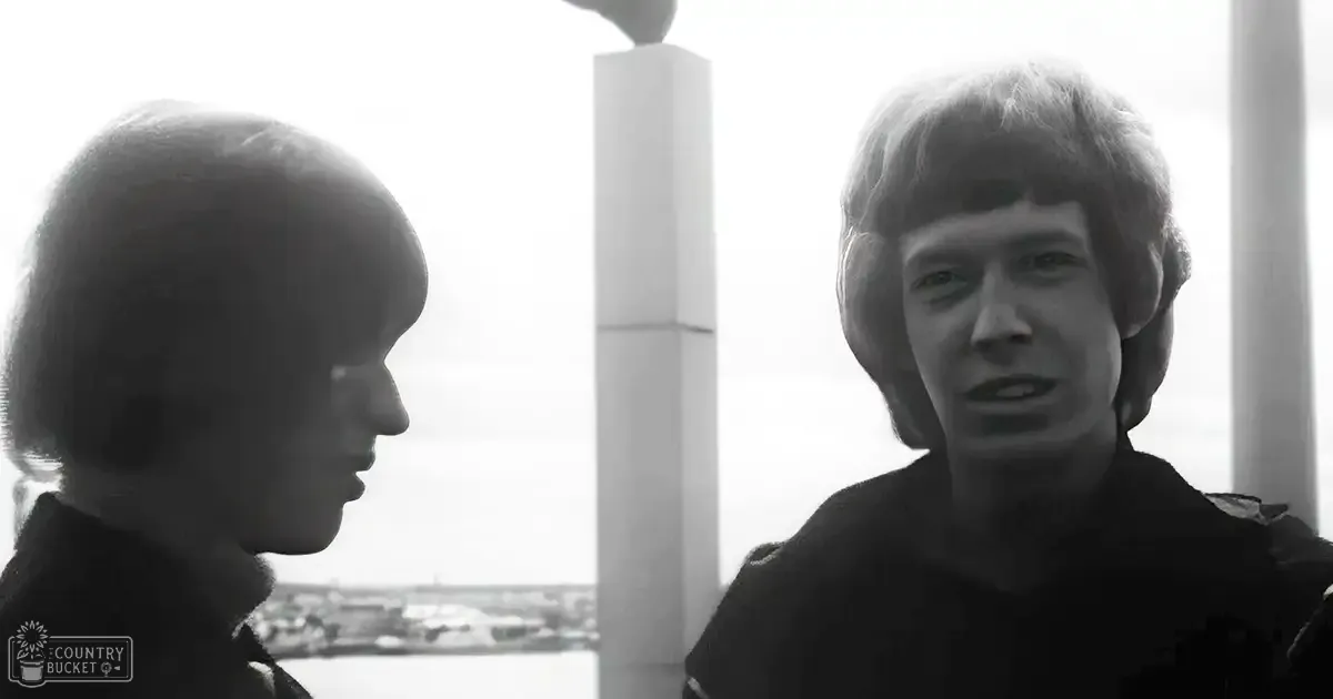 Two young adult males wearing dark, textured jackets with high collars stand by a waterfront at dusk, near a large concrete pillar.
