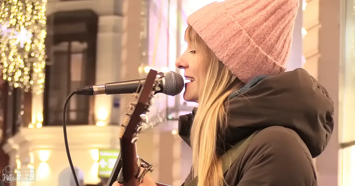 A young adult female with long blonde hair wearing a pink knit beanie, dark green hooded jacket, and possibly a scarf under it sings outdoors amidst holiday lights strung on buildings.