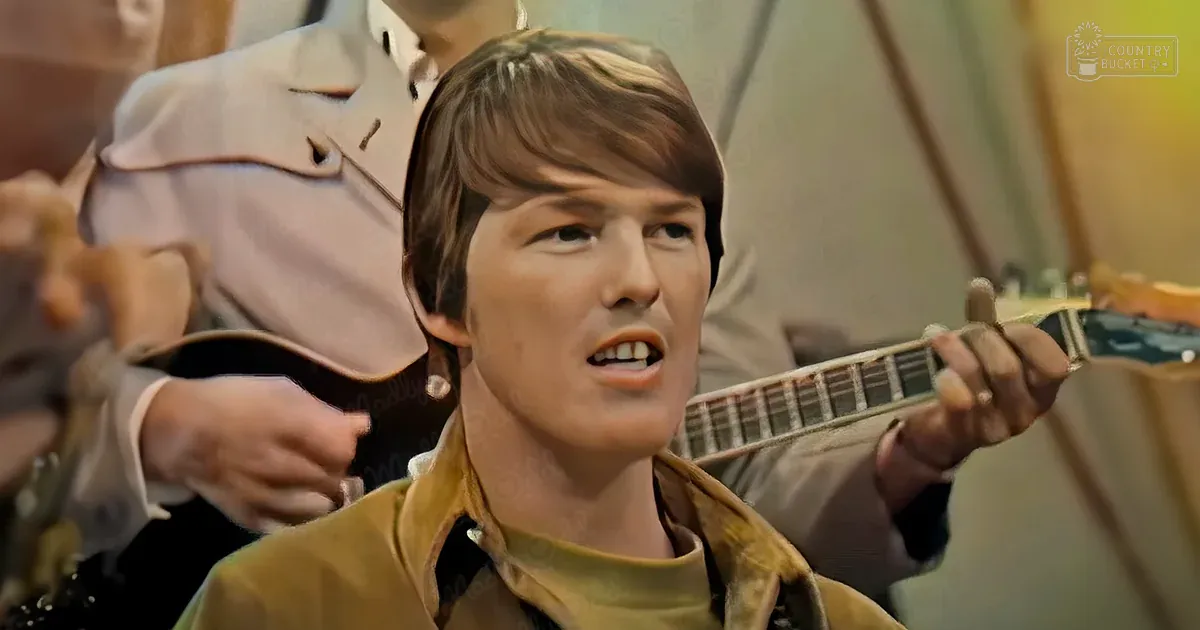 A young adult male plays a guitar in what seems to be an indoor setting with neutral-toned walls and possible furniture, suggesting a performance or recording session.