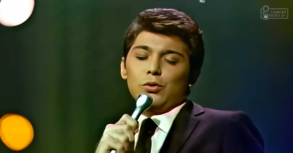 A young adult male in a dark suit jacket with a white shirt and dark tie, singing into a microphone on an indoor stage under warm-toned lighting.
