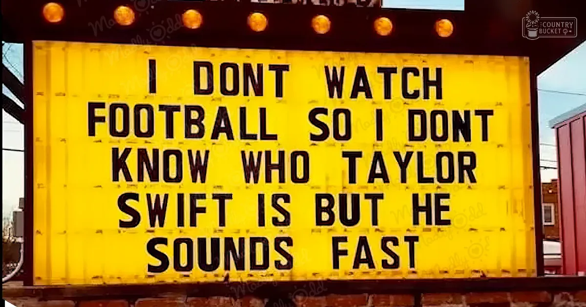 Yellow signboard with black text reading 'I DONT WATCH FOOTBALL SO I DON'T KNOW WHO TAYLOR SWIFT IS BUT HE SOUNDS FAST' mounted on a brick wall in an urban environment, with red and brown walls visible in the background.