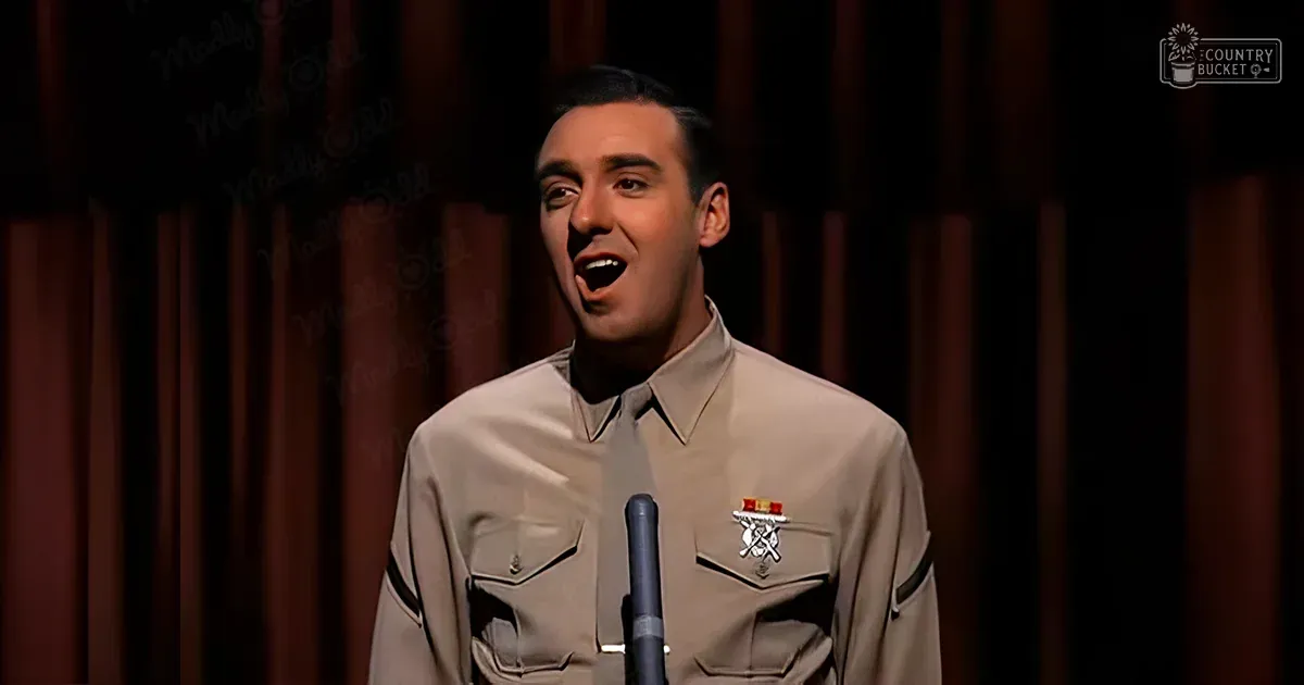 A young adult male in a light beige performance uniform with epaulets stands indoors, possibly on stage. A microphone is positioned directly in front of him.