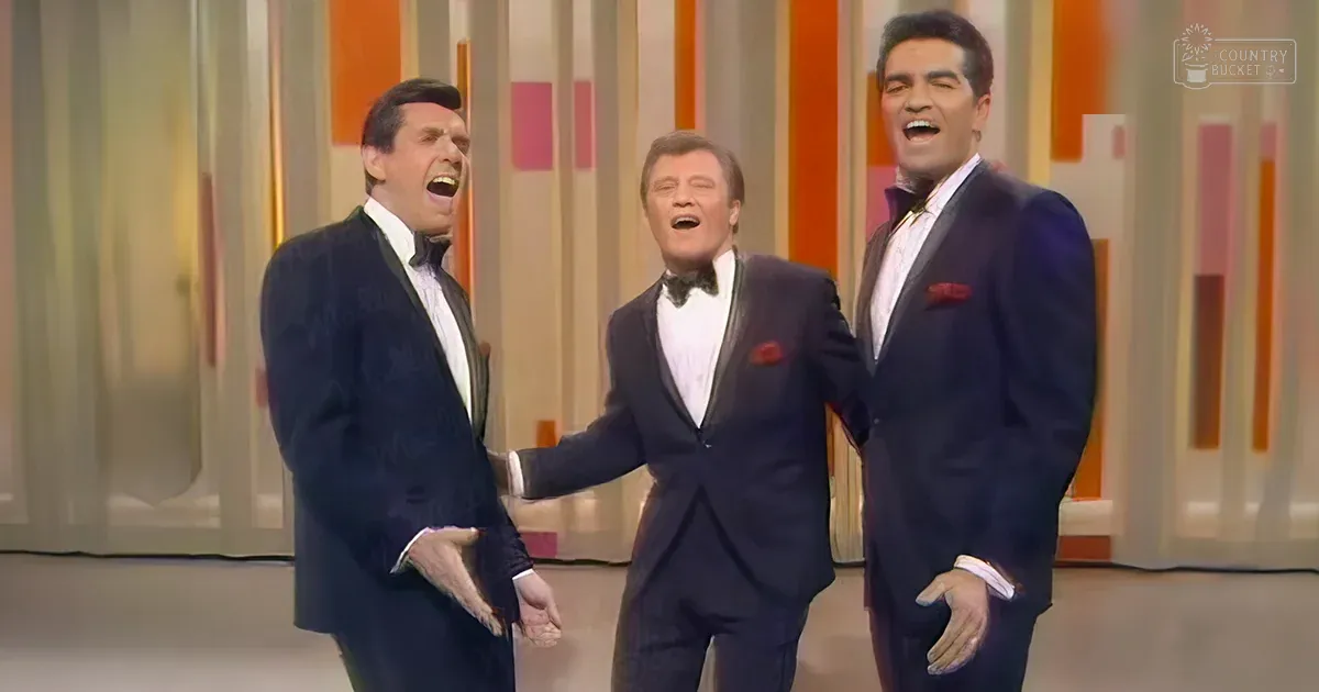 Group performance scene with three adult males in dark tuxedos on a brightly lit indoor stage with vibrant orange, pink, and white vertical striped backdrop.