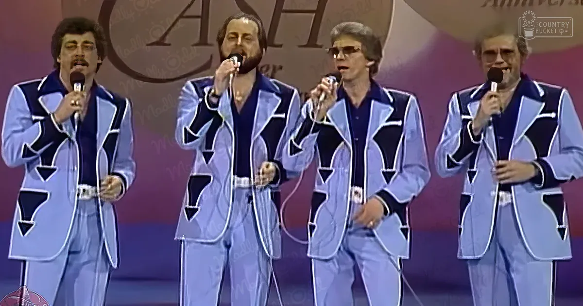 A musical performance group in 1970s attire performs on stage in an indoor venue, with microphones held by each member wearing light blue suits with dark blue accents.