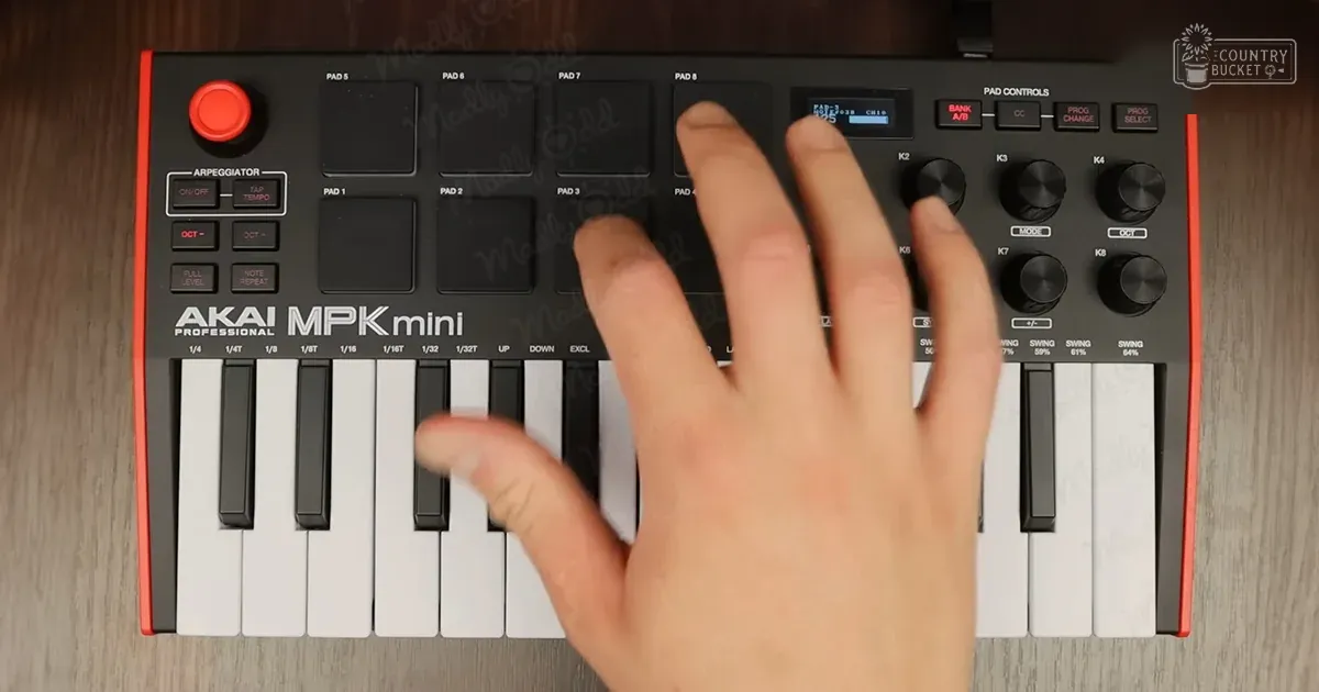 Close-up view of a hand interacting with an AKAI MPK Mini MIDI controller on a wooden surface, likely indoors.