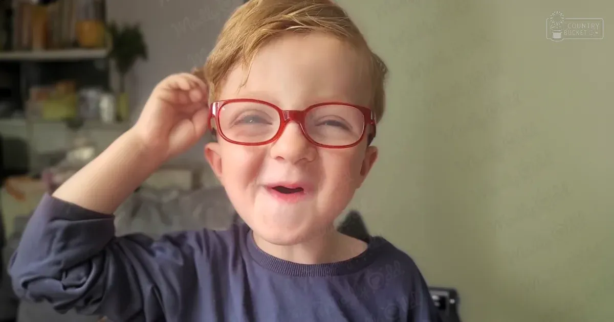 A toddler to young child wearing a dark blue, long-sleeved shirt with red-framed glasses indoors during daytime.