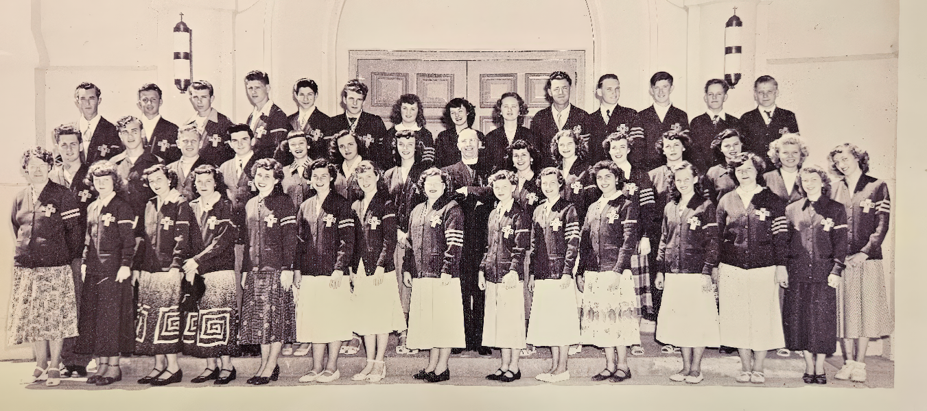 Indoors at a formal event with adults in blazers and skirts, and teens/young adults in dark jackets and cream-colored dresses with decorative patterns.