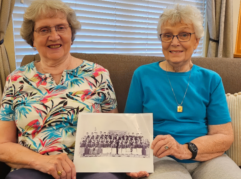 Two older adult females seated on a beige sofa in a living room with natural light filtering through horizontal blinds, wearing glasses and casual clothing.