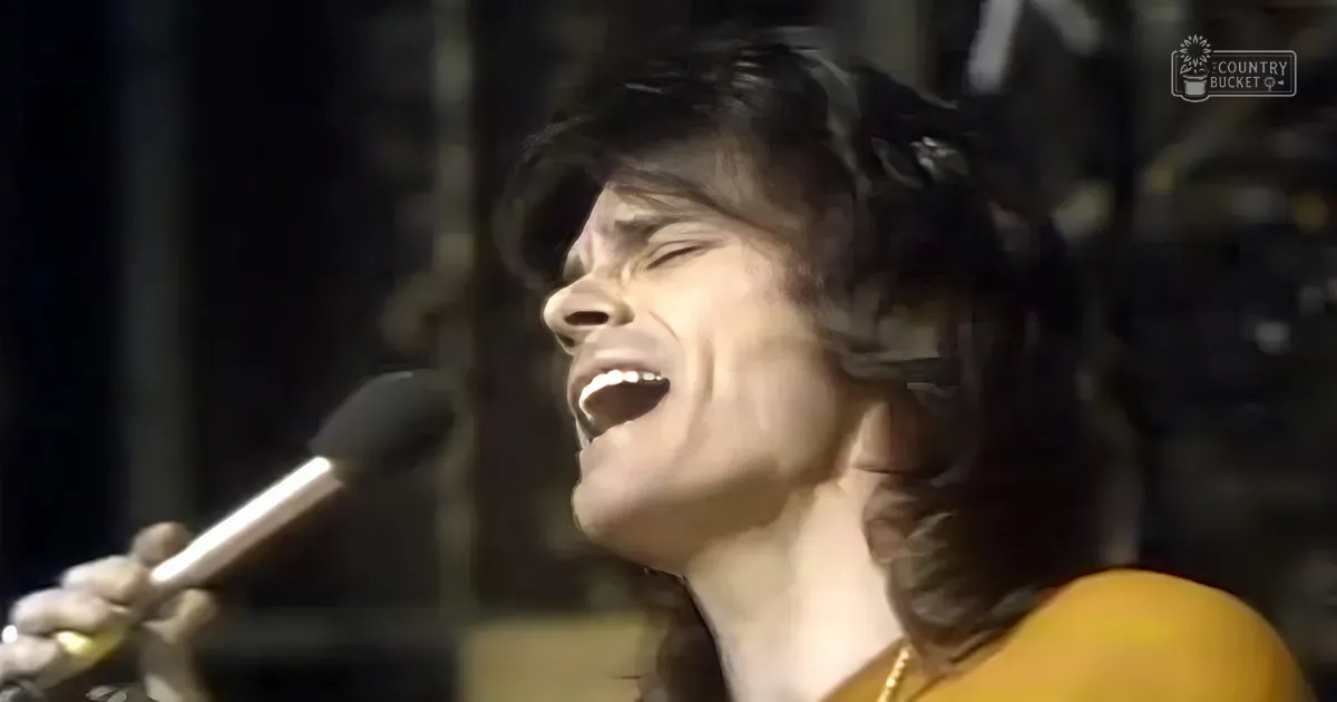 A young adult female singer wearing an orange, high-necked, long-sleeved garment with a gold necklace performs on stage in a concert setting with dark stage lighting and 'Madly Odd' text visible.