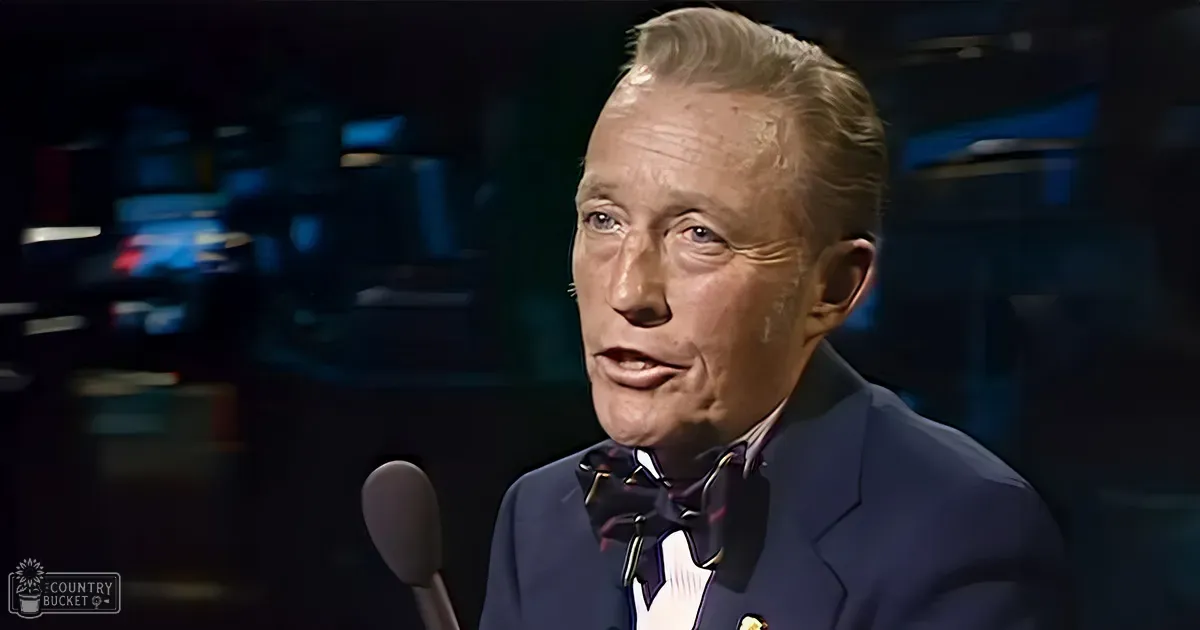 An older adult male in a studio setting, wearing a dark blue suit jacket with a bow tie and a collared shirt. The background suggests professional recording or broadcasting equipment.