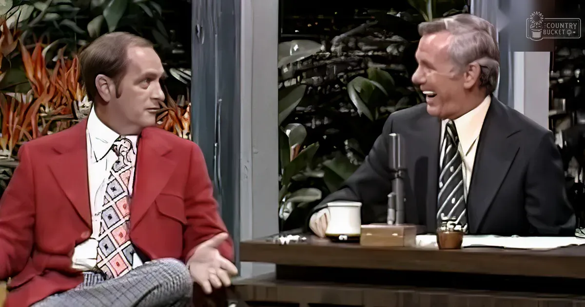 A talk show host in a red blazer with a white dress shirt and patterned tie, accompanied by an adult male guest in a black suit jacket and striped tie, seated at a wooden desk with studio plants in the background.
