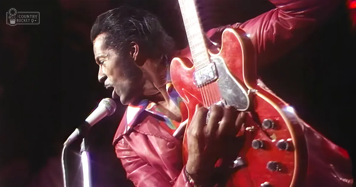 A male adult singer wearing a shiny red leather jacket performs on stage with a glossy red electric guitar and microphone stand, suggesting a retro or vintage style performance.
